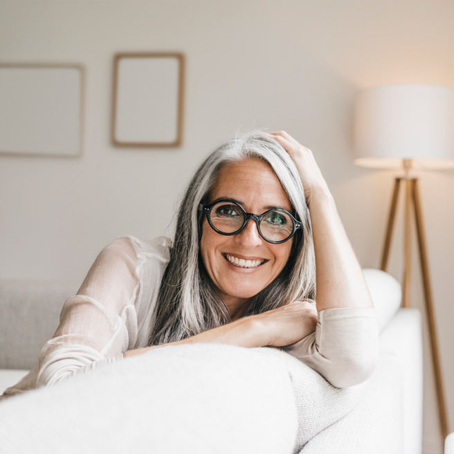 Eine lächelnde Frau mit grauen Haaren und einer runden Brille sitzt auf einem Sofa.