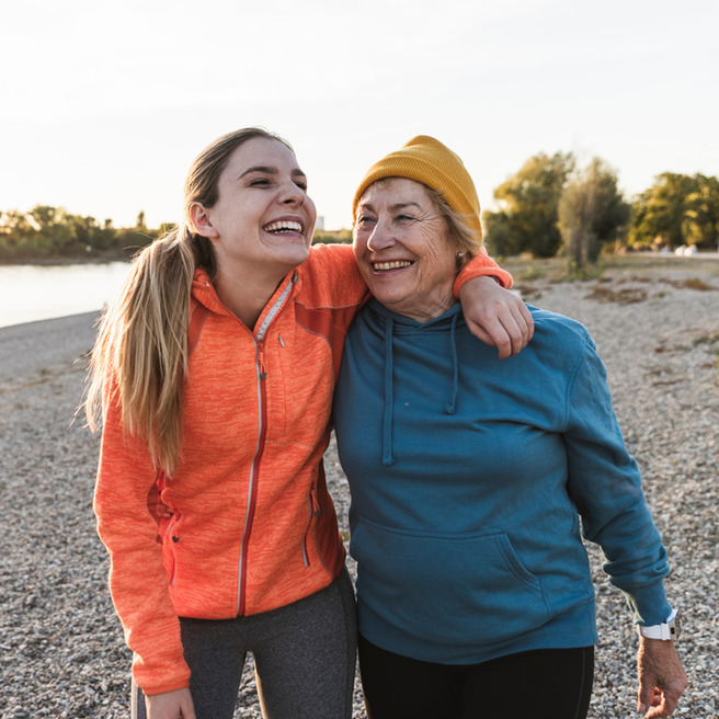 Zwei Frauen, eine junge und eine ältere, stehen eng nebeneinander am Ufer. Sie lächeln und haben Arme umeinander gelegt. Die jüngere Frau trägt eine orangefarbene Sportjacke, die ältere eine blaue Kapuzenjacke und eine gelbe Mütze. Beide wirken glücklich und entspannt.