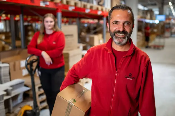 Ein lächelnder Mann in roter Arbeitskleidung hält ein Paket in der Hand, während eine Frau im Hintergrund ebenfalls in roter Kleidung steht. Beide arbeiten in einem Lager mit Regalen voller Kartons.