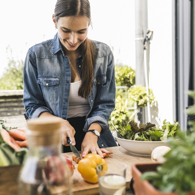 Eine Frau in einem Jeanshemd schneidet frisches Gemüse auf einem Holztisch. Vor ihr liegt eine bunte Auswahl an Gemüse und ein Schüssel mit Salat. Im Hintergrund sind Pflanzen zu sehen, die eine angenehme Atmosphäre schaffen.