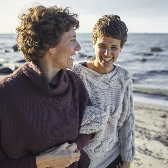 Zwei Frauen stehen am Strand, lachen und schauen sich an. Sie tragen gemütliche Pullover und haben kurze Haare. Im Hintergrund sind Wellen und Steine zu sehen, während die Sonne scheint.
