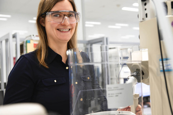 Eine lächelnde Frau in Schutzbrille steht an einem Laborgerät. Im Hintergrund sind Regale mit Laborutensilien zu sehen. Die Szene vermittelt eine positive und konzentrierte Arbeitsatmosphäre im wissenschaftlichen Umfeld.
