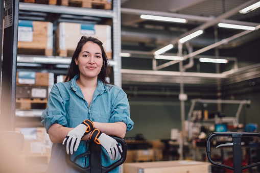 Eine Frau steht mit verschränkten Armen an einem Gabelstapler. Sie trägt Handschuhe und blickt freundlich in die Kamera. Im Hintergrund sind Holzpaletten und Regale sichtbar, die auf eine Lager- oder Werkstattumgebung hindeuten.