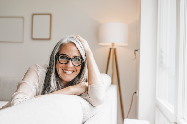 Eine lächelnde Frau mit grauen Haaren und einer runden Brille sitzt auf einem Sofa.